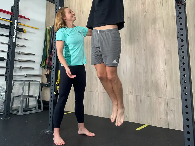 La sala de entrenamiento: el rack con la barra de dominadas, las barras y los discos, y una entrenadora junto a alguien que se cuelga de la barra