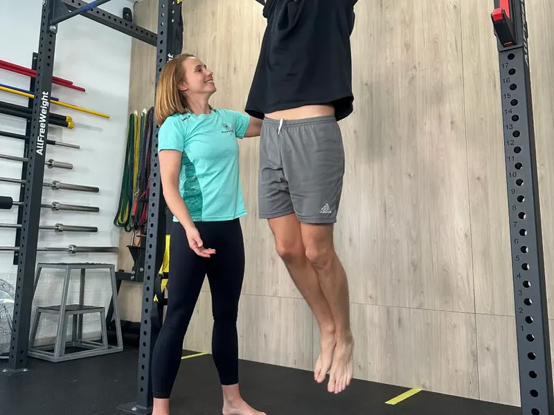 El rack de la sala de entrenamiento, con las barras y la pared de madera; una entrenadora acompaña a alguien colgado de la barra